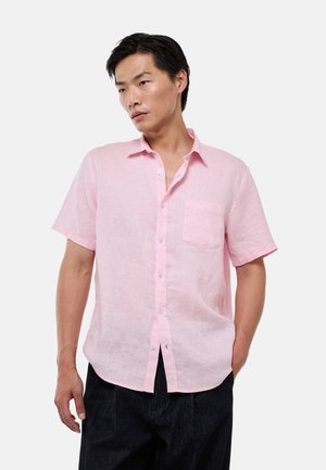 Homme portant une chemise rose clair à manches courtes avec une poche poitrine, associée à un pantalon sombre, debout devant un fond uni.
