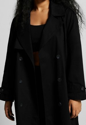 Manteau noir en trench avec un design à double boutonnage, de grands revers, un tissu texturé et des boutons noirs. Porté par-dessus un haut noir à côtes.