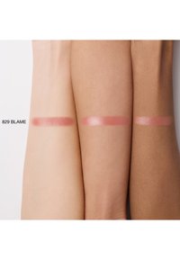 Des échantillons de la même teinte cosmétique, "829 Blame," sur trois bras de teintes de peau variées montrant une nuance rose pailletée.