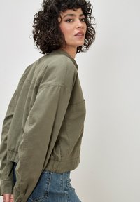 Olive green cotton bomber jacket with a relaxed fit, ribbed cuffs, and a single chest pocket, featured on a model against a neutral backdrop.
