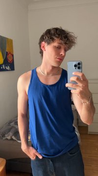 Giovane uomo con i capelli ricci che indossa una canotta blu e una collana d'argento si fa un selfie allo specchio con uno smartphone in una camera da letto.