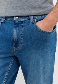 Denim džinsi vidēji zilā krāsā ar nolietotu apdari, ar rāvējslēdzēju, pogu priekšā un sānu kabatu. Izšūta detaļa ap kabatām.