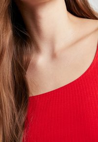 Top rojo con relieve y escote ancho, textura suave y un sutil brillo, combinado con cabello largo y castaño que cae sobre el hombro.