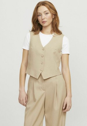 Beige Leinenweste mit V-Ausschnitt, fünfknöpfigem Frontverschluss und ärmellosem Design, kombiniert mit einem weißen T-Shirt und beigen Hosen.