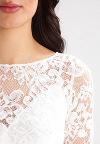 Vit spetsblus med blommönster, genomskinlig textur och rundad halsringning. Tyget är lätt och har intrikata detaljer.