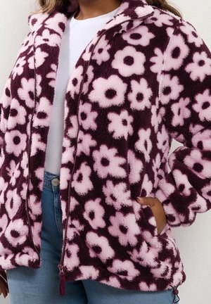 Kvinde iført en fleecejakke med lynlås med store pink blomster på en mørk lilla baggrund, kombineret med hvid skjorte og blå jeans.