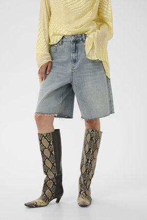Personne portant un short en denim clair jusqu'aux genoux, un haut jaune texturé à manches longues et des bottes montantes à motif peau de serpent.