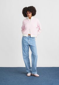 Chaqueta bomber de canalé rosa con acentos blancos, combinada con jeans de mezclilla claros a rayas y zapatos planos rosas. La modelo lleva gafas de sol moradas.