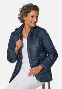 Veste matelassée marine avec une finition texturée, un motif matelassé et une capuche. Dotée d'un zipper frontal et de poignets élastiques. Portée par-dessus un haut blanc.