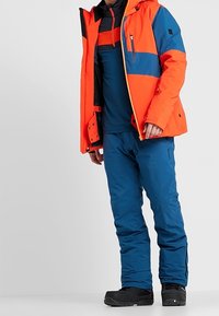 Giacca isolante arancione brillante e blu con cappuccio, abbinata a pantaloni blu e stivali da neve neri, dotata di cerniere e tasche.