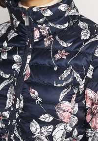 Veste matelassée bleu marine avec un motif floral comprenant des fleurs roses et blanches. Conçue avec un col montant et une fermeture éclair centrale.