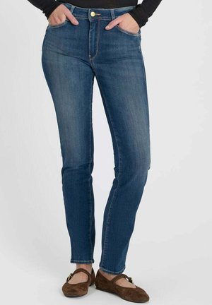 Vrouw draagt slim-fit blauwe spijkerbroek met handen in de zakken en bruine platte schoenen tegen een effen achtergrond.