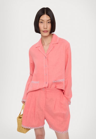 Set de cămașă și pantaloni scurți din in coral, cu guler, deschidere prin butoni și dantelă albă pe buzunare și tiv. Modelul ține o geantă țesută.