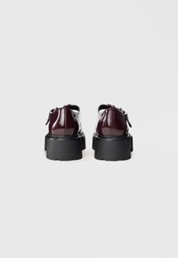Un par de zapatos de charol burdeos oscuros con suelas gruesas negras, vistos desde atrás sobre un fondo blanco liso.
