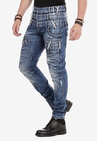 Blaue Jeans aus Denim haben ein robustes Design mit mehreren Taschen, abgenutzten Details und kontrastierendem weißen Nähten. Tragen Sie sie mit schwarzen Stiefeln.