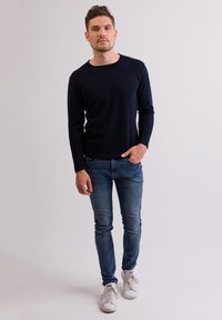 Granatowy sweter z dzianiny, dopasowany fason, okrągły dekolt, w połączeniu z wąskimi niebieskimi dżinsami i białymi trampkami, stojący na tle w neutralnym kolorze.