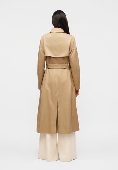 Γυναίκα με μακριά σκούρα μαλλιά που φοράει μπεζ καμπαρντίνα με ζώνη και κρεμ παντελόνι με φαρδιά γραμμή, στέκεται με την πλάτη γυρισμένη σε απλό φόντο.