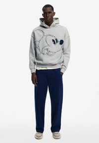 Sudadera gris claro con un gran bordado de la cara de un personaje de dibujos animados, combinada con pantalones de pierna ancha azul marino y zapatillas casuales beige.