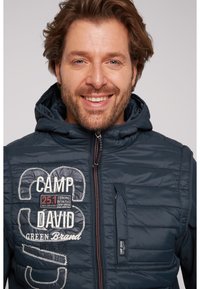 Vest puffer albastru închis cu glugă, având logo-uri și detalii cusute cu alb, fermoar frontal și țesătură texturată pe stratul exterior.