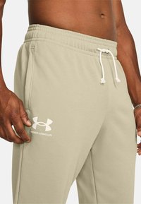 Pantalones de chándal color beige claro con una cintura elástica y cordón blanco. Cuenta con bolsillos laterales y un logo blanco de Under Armour en el muslo.