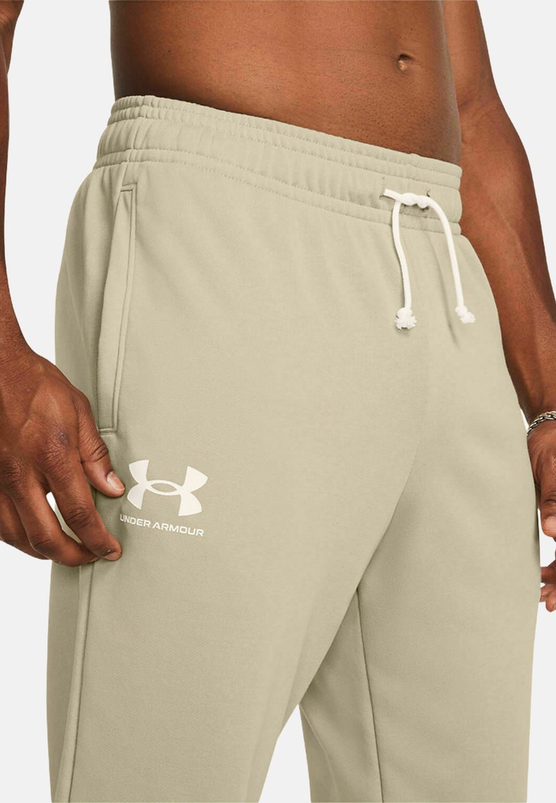 Jasno beżowe dresy z elastycznym pasem i białym sznurkiem. Posiadają boczne kieszenie oraz białe logo Under Armour na udzie.