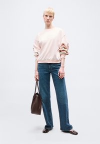 Γυναίκα με κοντά ξανθά μαλλιά, φοράει ανοιχτό ροζ φούτερ με μανίκια με floral μοτίβο, μπλε τζιν, σκούρα καφέ παπούτσια και κρατάει καφέ τσάντα χειρός.