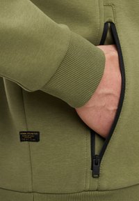 Olijfgroene sweatshirt met geribde manchetten, voorzien van een zwarte ritszak en een klein zwart label dat aan de zoom is gestikt. Gladde stoftextuur.