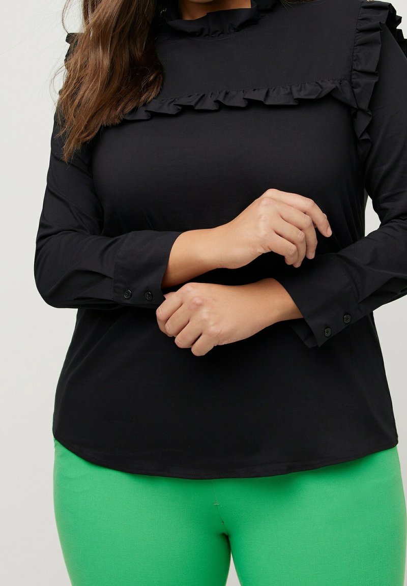 Haut noir à manches longues avec des détails en volants le long de la poitrine et du col, avec des poignets à boutons. Associé à un pantalon ajusté vert vif.