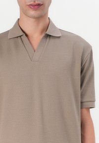 Béžové polo tričko s texturovaným vzorem ve tvaru včelí plástve. Má výstřih do V a krátké rukávy, vyrobené z prodyšného materiálu.