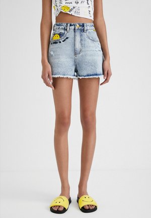Person wearing light blue denim shorts with smiley face embroidery and yellow smiley face slides on feet.