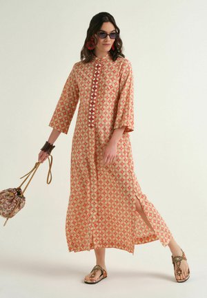 Donna che indossa un abito lungo arancione e beige a motivo con spacco frontale, sandali, orecchini rossi, occhiali da sole e che tiene una piccola borsa fantasia paisley.