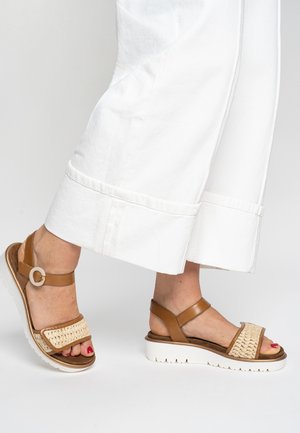 Brune lædersandaler med vævet tekstildetalje, justerbar ankelrem og hvid skridsikker sål. Viser fodplacering ved siden af et hvidt bukseben.