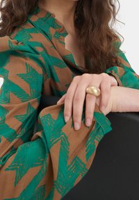 Blusa marrone e verde con motivo e colletto a balze. Una mano si appoggia su una superficie scura, indossando un anello d'oro texture.