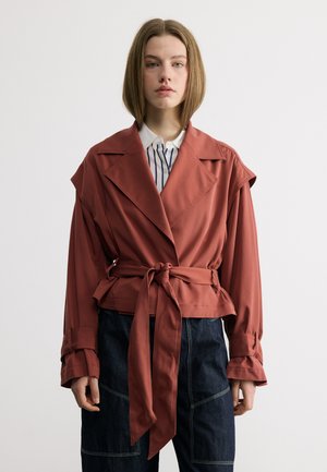 Mujer joven con chaqueta color óxido ceñida con cinturón sobre una camisa blanca con rayas negras, combinada con pantalones vaqueros oscuros, de pie frente a un fondo blanco.