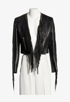 Chaqueta de cuero negra recortada con flecos largos, ojales dorados y bolsillos con cremallera, usada sobre pantalones blancos de talle alto en un maniquí.