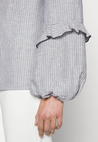 Blouse rayée à poignets volantés, présentant de fines rayures verticales bleu marine et blanches. Le tissu a une texture douce, avec un design de manches bouffantes.