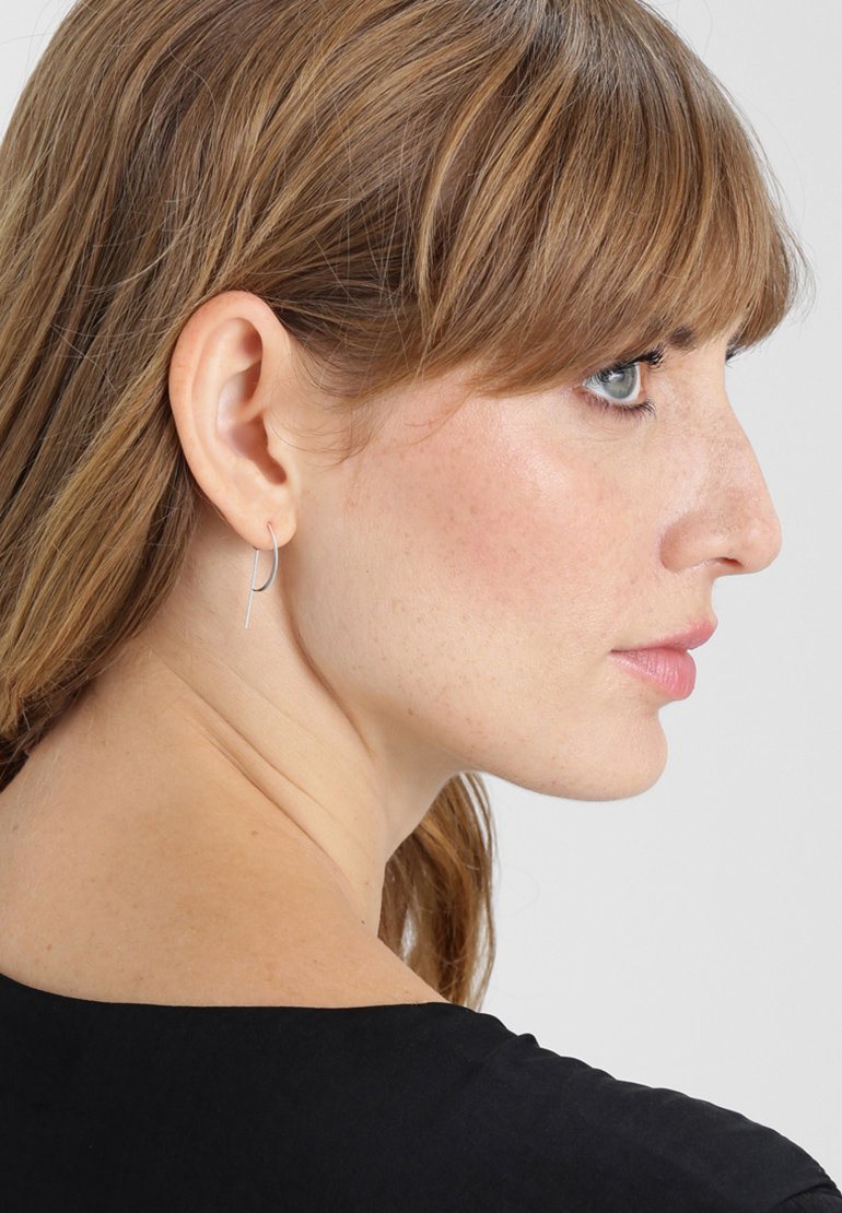 Pendientes de aro de plata con un diseño minimalista, que presentan una forma de alambre delgado y un acabado pulido, llevados por una persona con una blusa negra.