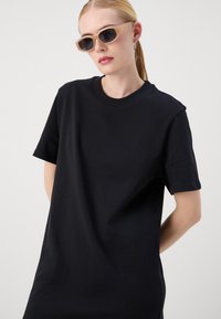 Black cotton t-shirt with short sleeves and a round neckline. Model wears light-colored sunglasses with rectangular frames.