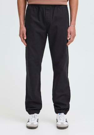 Black elastic-waist trousers with ankle cuffs, featuring a smooth texture and straight leg design, paired with white and grey sneakers.