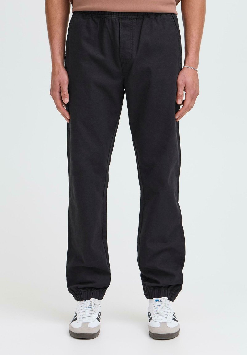 Schwarze Hose mit elastischem Bund und Knöchelbündchen, mit glatter Textur und geradem Bein, kombiniert mit weißen und grauen Sneakern.