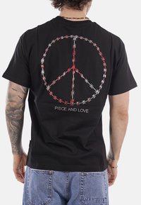 Uomo con maglietta nera con simbolo di pace a filo spinato rosso e bianco e scritta "Piece and Love", abbinata a jeans blu e tatuaggi sulle braccia.