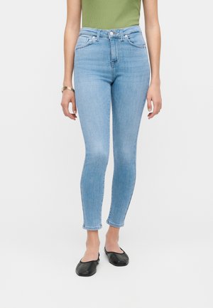 Kvinde iført lyseblå skinny jeans med høj talje, grøn ærmeløs top, guldarmbånd og sorte flade sko står mod en hvid baggrund.
