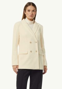 Doppelreiher-Blazer in Creme mit geripptem Muster, zwei Fronttaschen und hellen Knöpfen. Tragend über einem Rollkragenpullover. Maßgeschneiderte Form mit Revers.