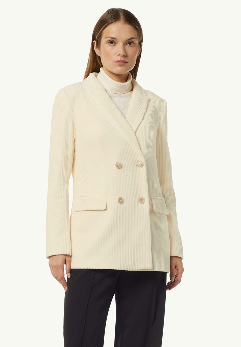 Doppelreiher-Blazer in Creme mit geripptem Muster, zwei Fronttaschen und hellen Knöpfen. Tragend über einem Rollkragenpullover. Maßgeschneiderte Form mit Revers.
