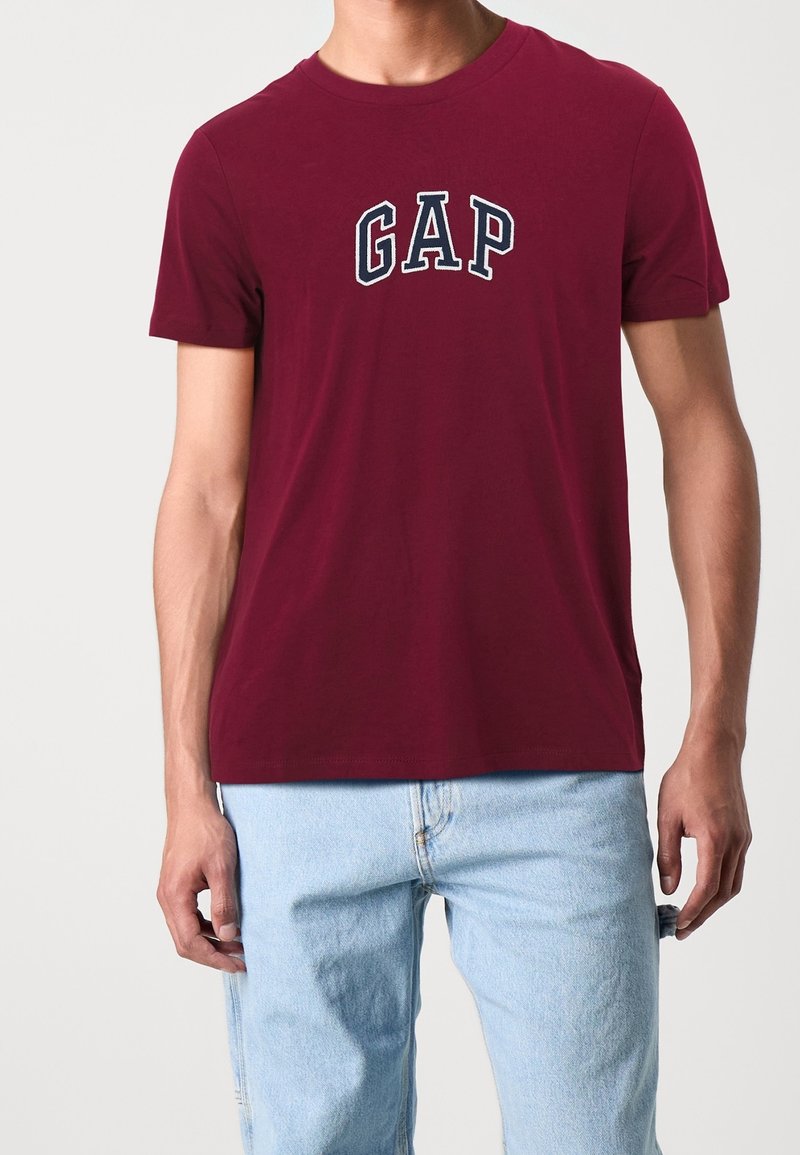Bordowy bawełniany T-shirt z białym, haftowanym logo „GAP” na piersi. Regularny krój, krótkie rękawy i okrągły dekolt.