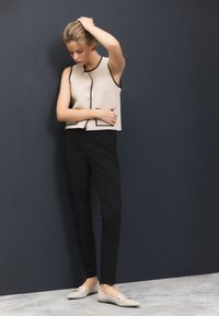 Ujjatlan bézs felső fekete szegéllyel és gombokkal, hozzáillő fekete nadrággal és világosszürke bebújós cipőkkel. Minden anyag sima textúrájú.