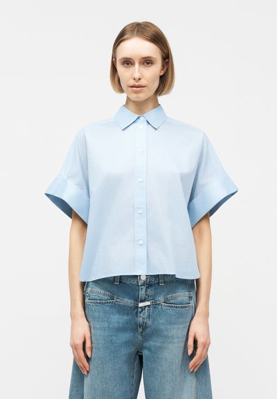 Femme aux cheveux blonds courts portant une chemise à manches courtes bleu clair boutonnée et un jean bleu ample, posant devant un fond uni.