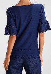 Donna che indossa un top blu navy con maniche e orlo in pizzo traforato a forma di scalloped, abbinato a jeans blu scuro, vista da dietro.