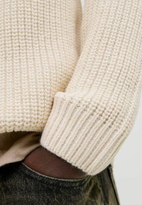 Pull en tricot couleur crème avec un motif texturé et des poignets côtelés, associé à un denim vert foncé. Accent sur le tissu et les détails.