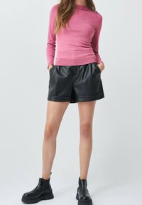 Kobieta w różowej bluzce z długim rękawem, czarnych szortach z sztucznej skóry i czarnych, masywnych botkach na kostce stoi na tle jednokolorowym.